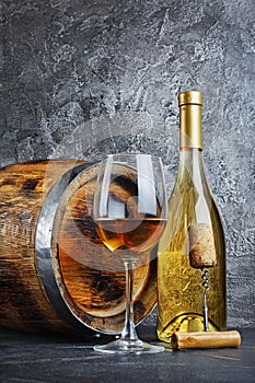 White wine bottle with glass for tasting and wooden barrel with corkscrew in dark cellar