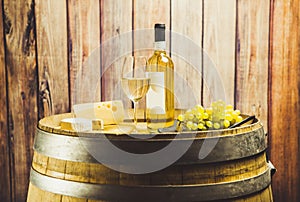 White wine in glass, bottle, grape and cheese on barrel front wood background.