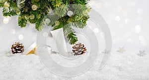 White winter skin care cosmetic products in snow with Christmas tree, pine cones and bokeh lights. Opened face cream jar