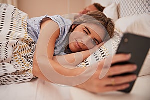 White woman half asleep in bed looking at smartphone, her partner sleeping in background, close up