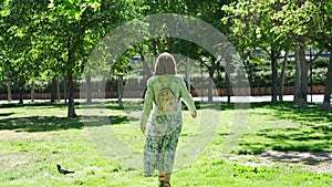 White woman with her back walking through a sunny public park with plants and trees.