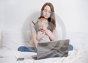 White woman sitting on a bed with a small child, working at a laptop ,  young mother working at home