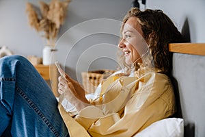 White woman using cellphone with earphones lying in bed at home