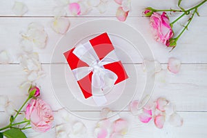 On a white wooden background there are roses and petals forming a circle, inside there is a gift in a red box and a