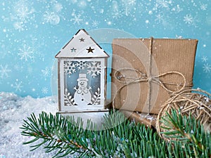 White wooden candle lantern with snowman next to handcrafted gift and twine ball over snowy fir tree branch.