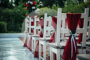 White wooden chairs decorated with red fabric and ribbons for wedding reception outdoor. Guest chairs in rows in the summer park,