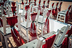 White wooden chairs decorated with red fabric and ribbons for wedding registration outdoor. Guest chairs in rows, close up