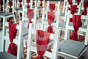 White wooden chairs decorated with red fabric and ribbons for wedding registration outdoor. Guest chairs in rows, close up