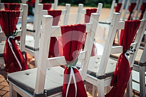 White wooden chairs decorated with red fabric and ribbons for wedding registration outdoor. Guest chairs in rows, close up