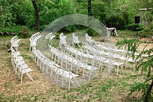 White wooden chairs placed in rows on the lawn. Venue of wedding ceremony or other events in botanic garden. Seats for visitors