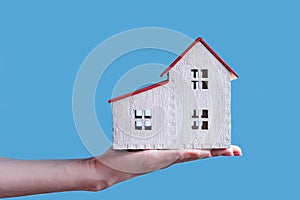 White wooden house on a female hand. Blue background. Housing concept