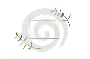 White wooden photo frame and green eucalyptus leaves on white background. Flat lay top view copy space. Stylish minimal