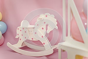 White wooden rocking horse with  stars and colorful balloons on pink background. Little girl room interior or first birthday party