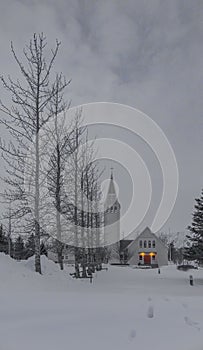 White wooden Selfosskirkja church with its gardens and walkways completely covered in snow under a light fog with a cloudy sky and