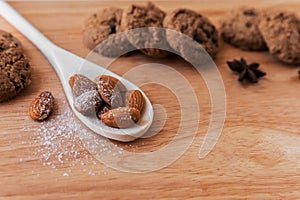 White wooden spoon with almonds, sprinkled with icing sugar on top, against a background of cookies with chocolate and star anise