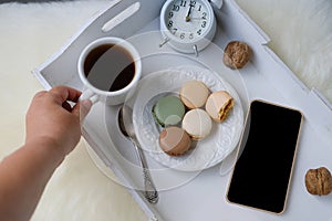 White wooden tray with black tea, coffee, plate with macaroon cakes, alarm clock, blank screen phone, concept of technology, time