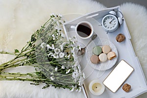 White wooden tray with black tea, coffee, plate with macaroon cakes, alarm clock, flowers, blank screen phone, concept of