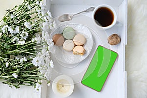 White wooden tray with black tea, coffee, plate with macaroon cakes, flowers, candles, blank screen phone, concept of technology,