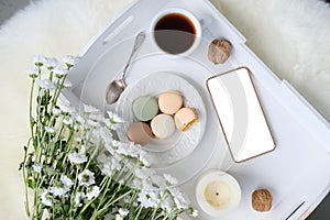 White wooden tray with black tea, coffee, plate with macaroon cakes, flowers, candles, blank screen phone, concept of technology,
