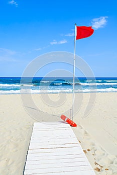 White wooden walkway on sandy beach with red flag on a pole, Porto Giunco bay, Sardinia island, Italy