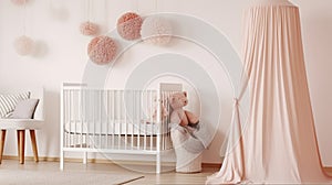 White woolen blanket and pastel pink pompom placed on a wooden crib with canopy in bright baby room interior with poster on the