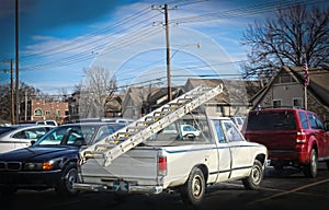 White work truck with plastic covered window and ladders tied to back parked in parking lot with houses in background