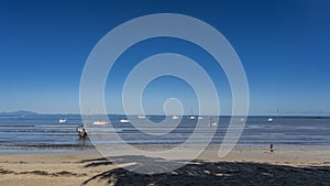 White yachts are visible in the blue ocean. A wooden fishing boat with a mast is moored in shallow water. The waves roll towards t