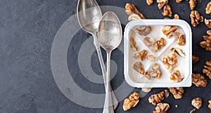 White yogurt in plastic square cup with marmalade and spoon in with walnuts on gray background. Flat view