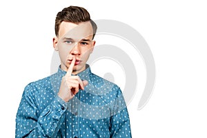 White young guy dressed in blue shirt showing silence gesture with his forefinger by mouth. Isolated on white background