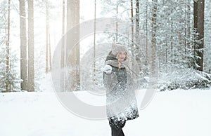 White young woman in a hat and mittens and a black jacket throwing snow in winter in the forest among the trees