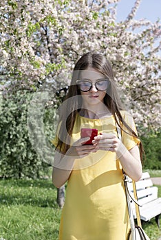 Young woman with long hair in a yellow dress with a phone on the background of a blooming garden