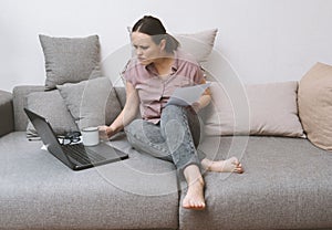 white young woman in pink blouse and gray jeans sitting on the couch with a laptop and a document in his hands and sullenly photo