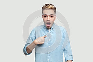 Who? me? Portrait of shocked handsome young man in light blue shirt standing, pointing himself and looking at camera with