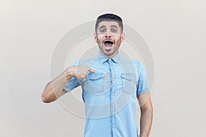 Who? me? Portrait of surprised handsome young bearded man in blue shirt standing, pointing himself and looking at camera with