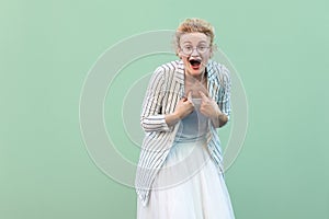 Who? me? Portrait of surprised young blonde woman in white striped blouse with eyeglasses standing, amazed face, pointing herself