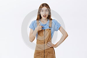 Who me. Surprised and stunned beautiful young girl in brown dungarees and blue t-shirt pointing at chest and staring