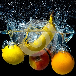 Whole banana, orange, lemon and apple in water on a black background. Fresh fruit with water spray