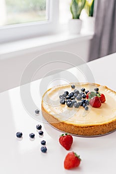 Whole cheesecake with fresh strawberries and blueberries on a white table in white modern kitchen.