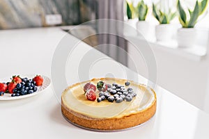 Whole cheesecake with fresh strawberries and blueberries on a white table in white modern kitchen.