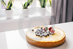 Whole cheesecake with fresh strawberries and blueberries on a white table in white modern kitchen.