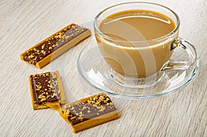 Whole cookie with caramel, cocoa and cereals, halves of cookie, cup of coffee with milk on saucer on wooden table