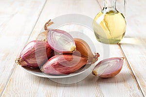 Whole and halved red shallots on a plate over white wooden table. Unpeeled eschalot bulbs close-up. Fresh raw long onion ready for