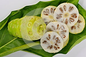 Whole Noni fruit Morinda citrifolia and slices cut crosswise, on leaf, against white background