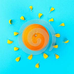 A whole orange in the form of the sun and rays from the petals of a yellow flower on a pastel blue background.