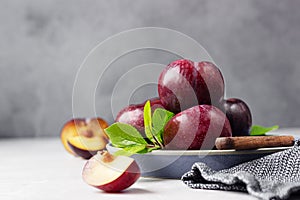 Whole purple plums and slices with leaves and knife on ceramic plate, light grey concrete background