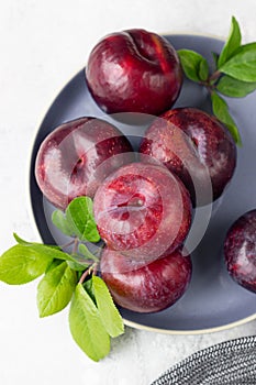 Whole purple plums and slices with leaves and knife on ceramic plate, light grey concrete background. Top view