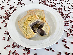 A whole and a quarter-cut mooncakes in a white round plate on white marble background with rd beans surrounding.