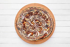 Whole round pizza on wooden plate topped with mushrooms, onion and chopped meat. Top view on white board background