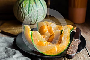 Whole and sliced ripe organic cantaloupe with juicy orange pulp, dark plate, knife, wood cutting board, rustic kitchen interior