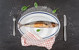 Whole smoked mackerel fish on white oval plate, gray black stone like table under, rock salt and green leaves salad near